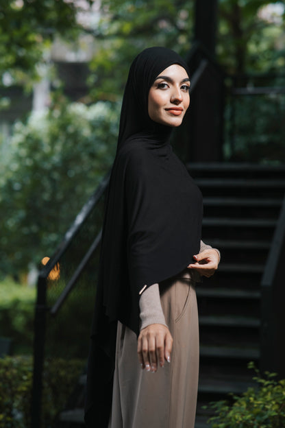 A woman wearing a soft black bamboo hijab and a dress stands outdoors in front of a staircase with green foliage in the background.