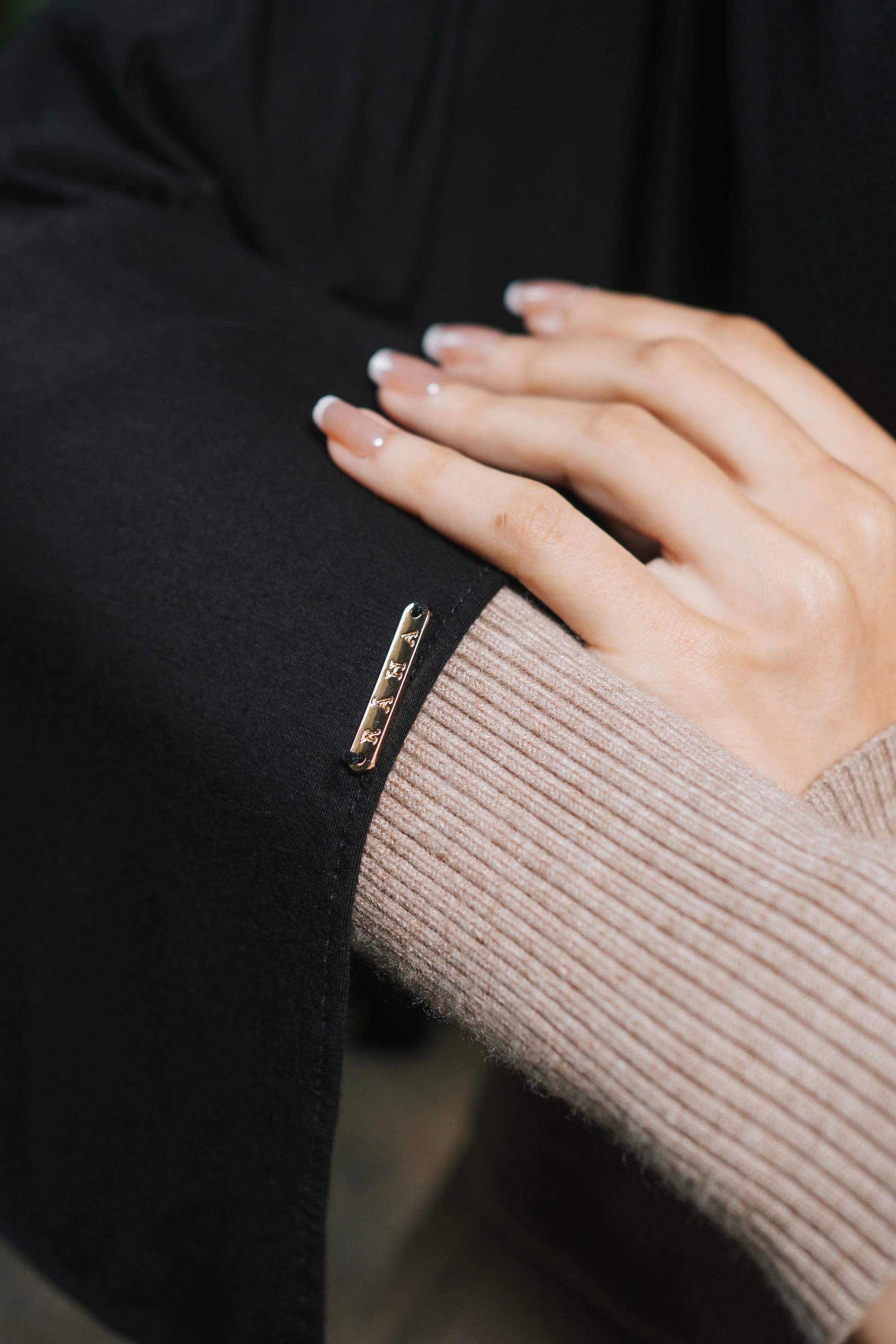 A close-up of a person's hand resting on a black draped bamboo hijab over the sleeve, featuring a small gold-toned rectangular badge that is engraved with the brand Raha.