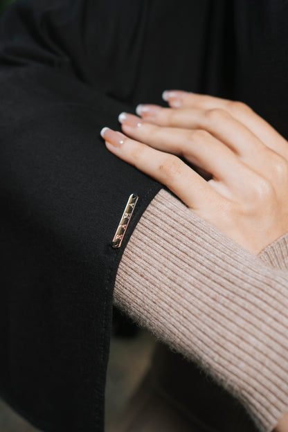 A close-up of a person's hand resting on a black draped bamboo hijab over the sleeve, featuring a small gold-toned rectangular badge that is engraved with the brand Raha.